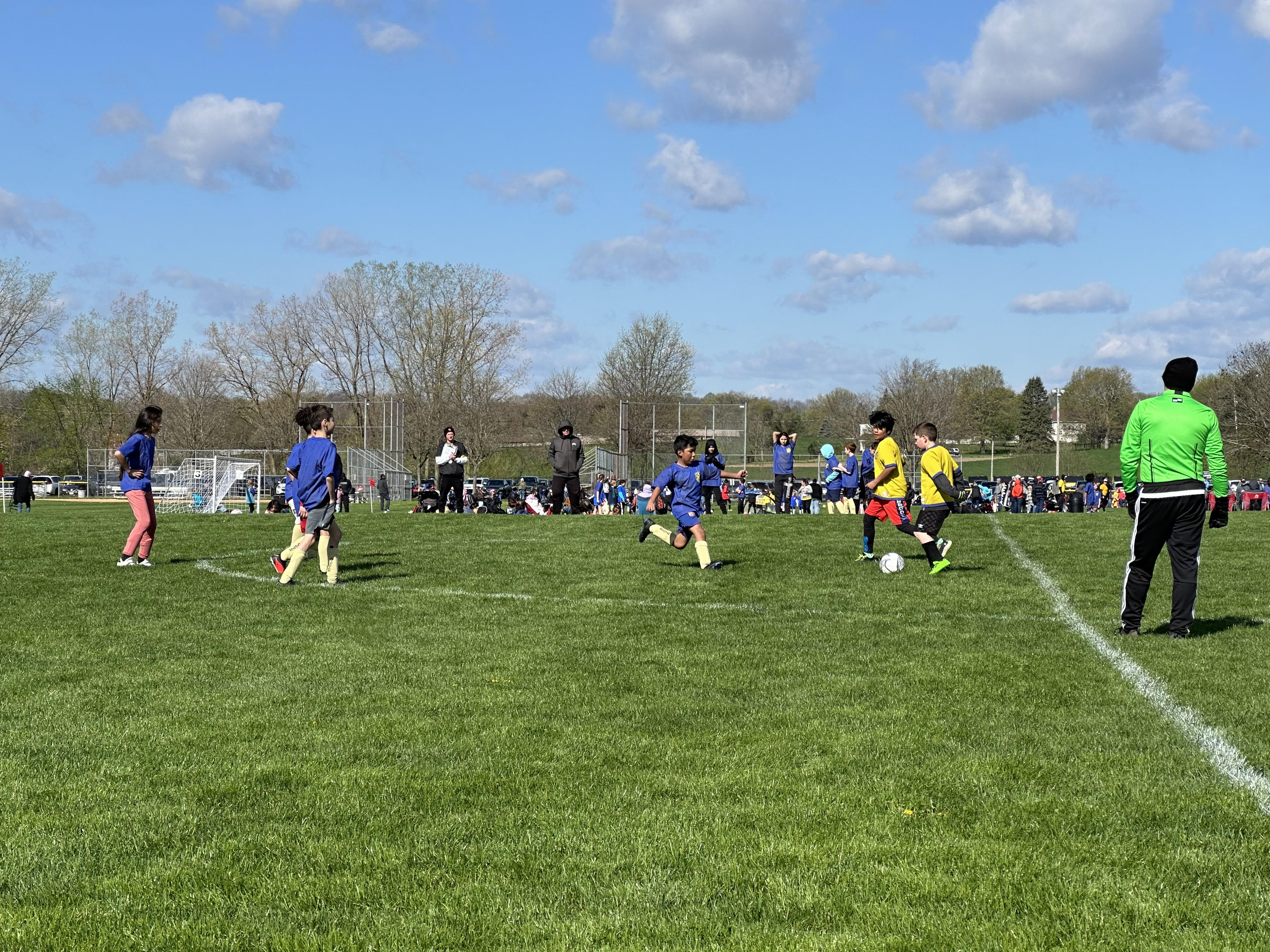 Children playing soccer on a grassy field, with a mix of participants wearing blue and yellow jerseys, surrounded by spectators in the background.